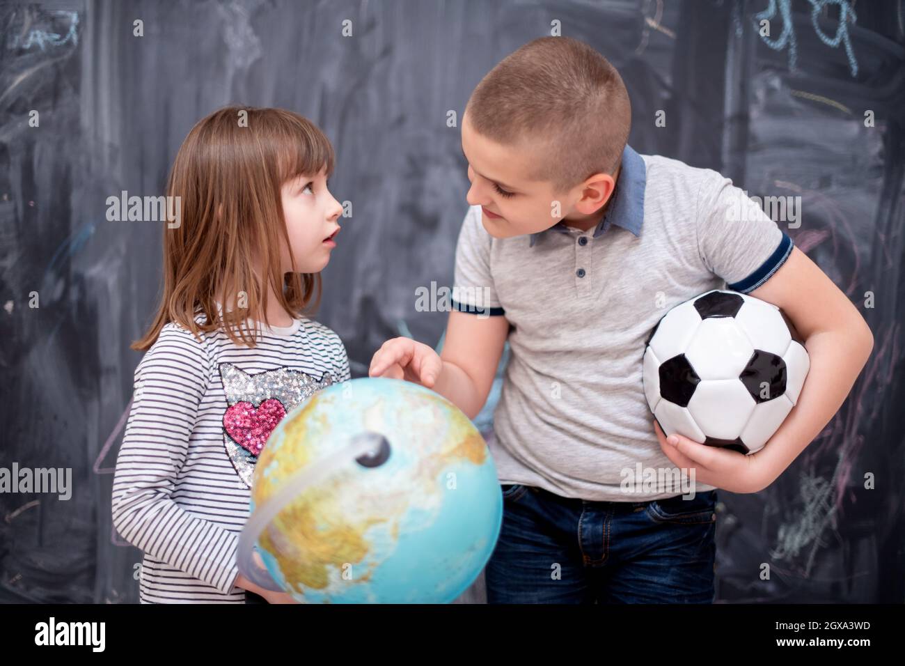 happy childrens boy with soccer ball and little girl learning about the world using globe of earth while standing in front of black chalkboard Stock Photo