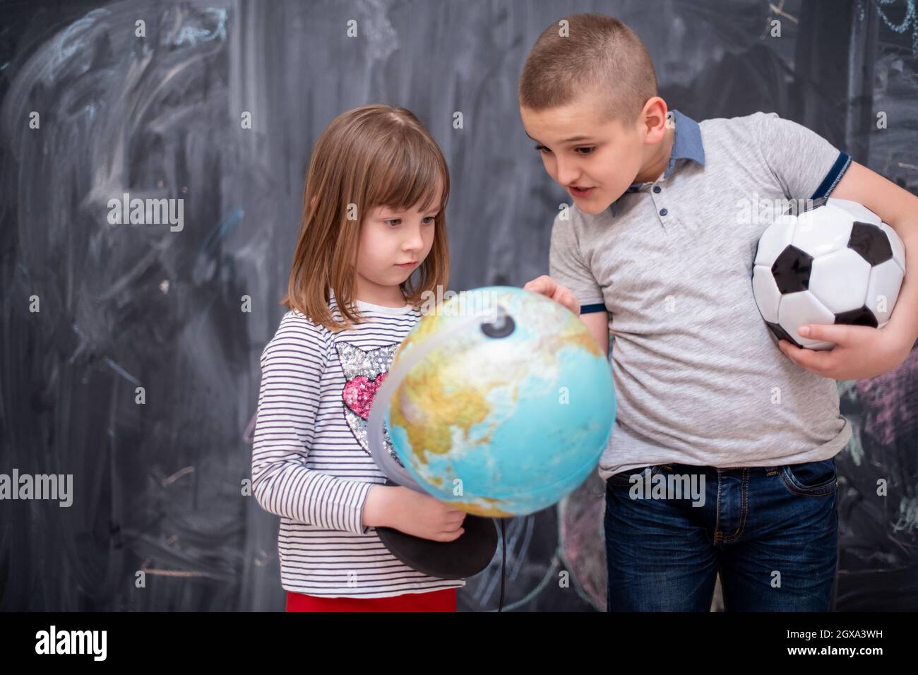 happy childrens boy with soccer ball and little girl learning about the world using globe of earth while standing in front of black chalkboard Stock Photo