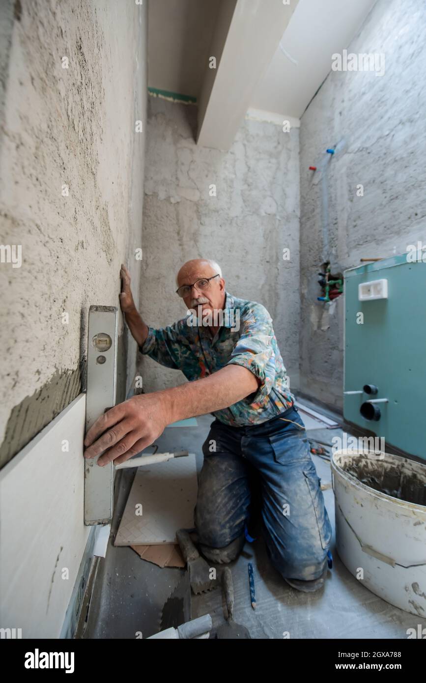 experienced senior construction worker using level tool while installing big ceramic tiles on interior concrete walls Stock Photo
