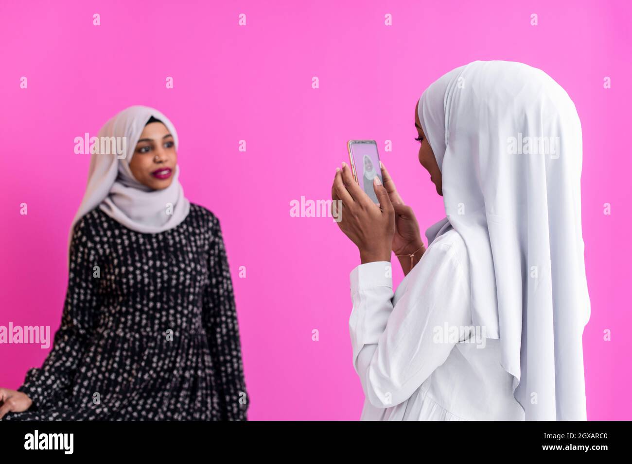 portrait of two diverse african student girls wearing scarf modern muslim clothes  taking self portrait with phone against plastic pink background Stock Photo