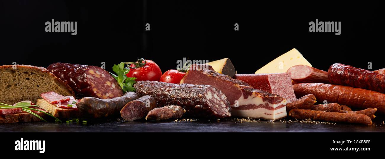 Assorted types of cured sausages neat bread placed on dark surface with vegetable and cheese in studio on black background Stock Photo