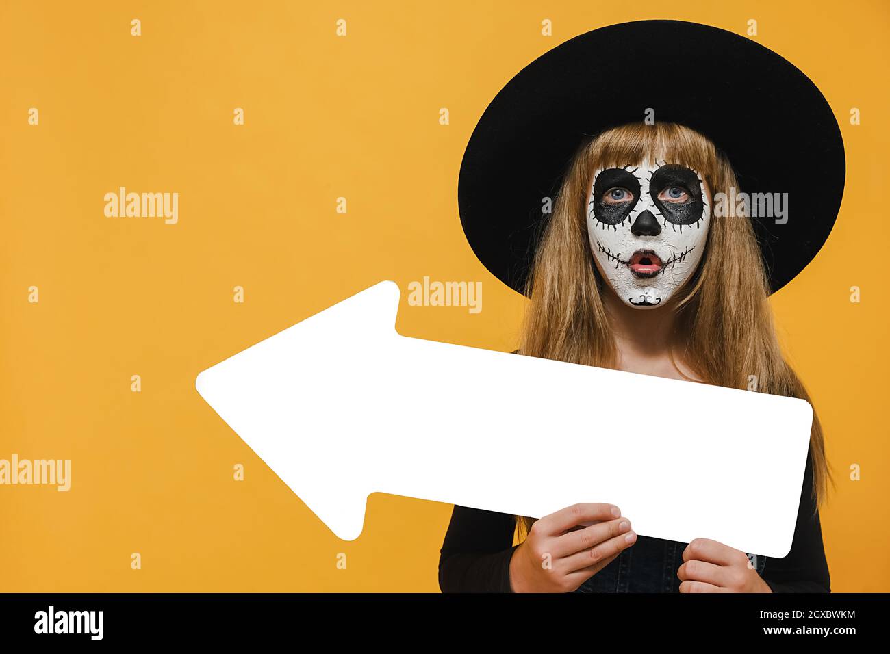 Scary bright alive girl child with Halloween makeup mask showing aside with white arrow wears big black hat, posing isolated over yellow color wall Stock Photo