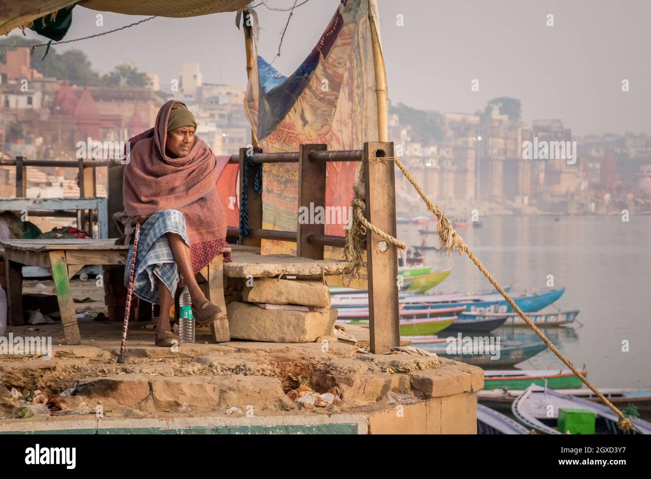 INDIA, VARANASI - NOVEMBER 27, 2015: Ethnic male sitting on wooden bench near boat on river embankment in India Stock Photo