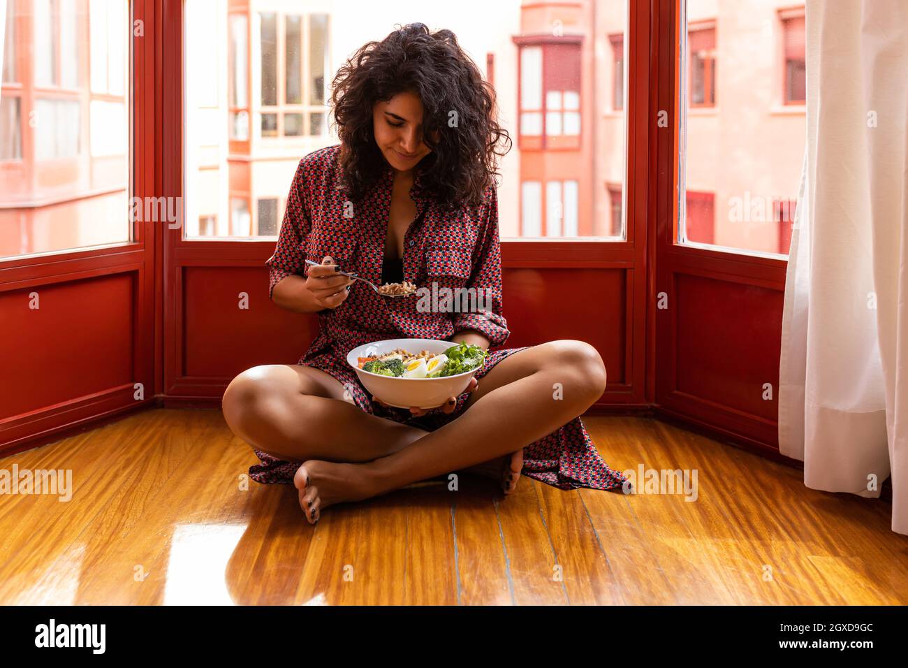 Young female with curly hair sitting with crossed legs and eating super food bowl with chickpea chopped eggs broccoli Stock Photo