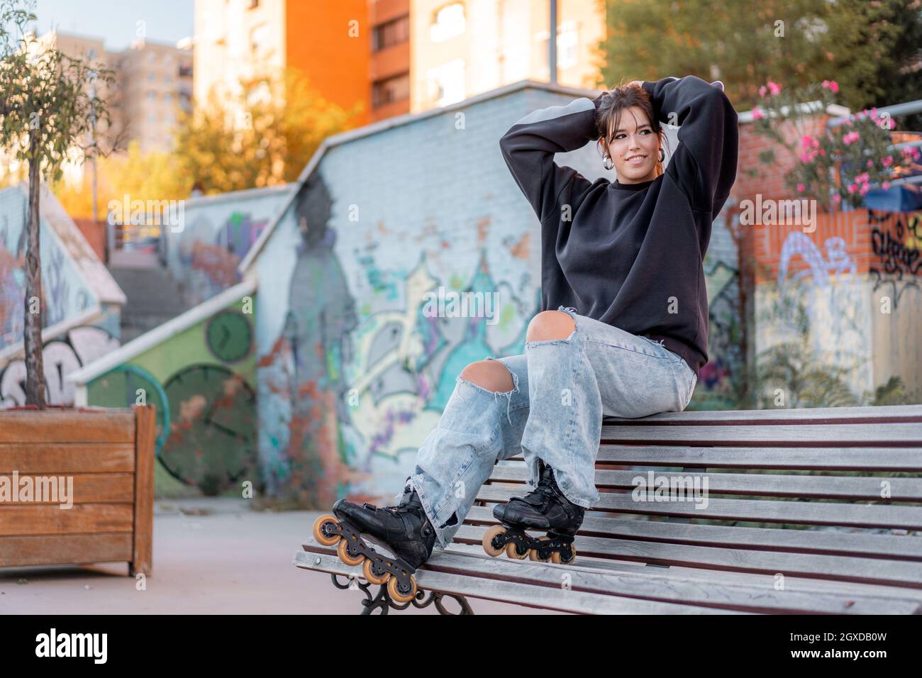 Full body of positive young female wearing casual black hoodie and light blue  jeans with slits and roller blades sitting on back of wooden bench in sk  Stock Photo - Alamy
