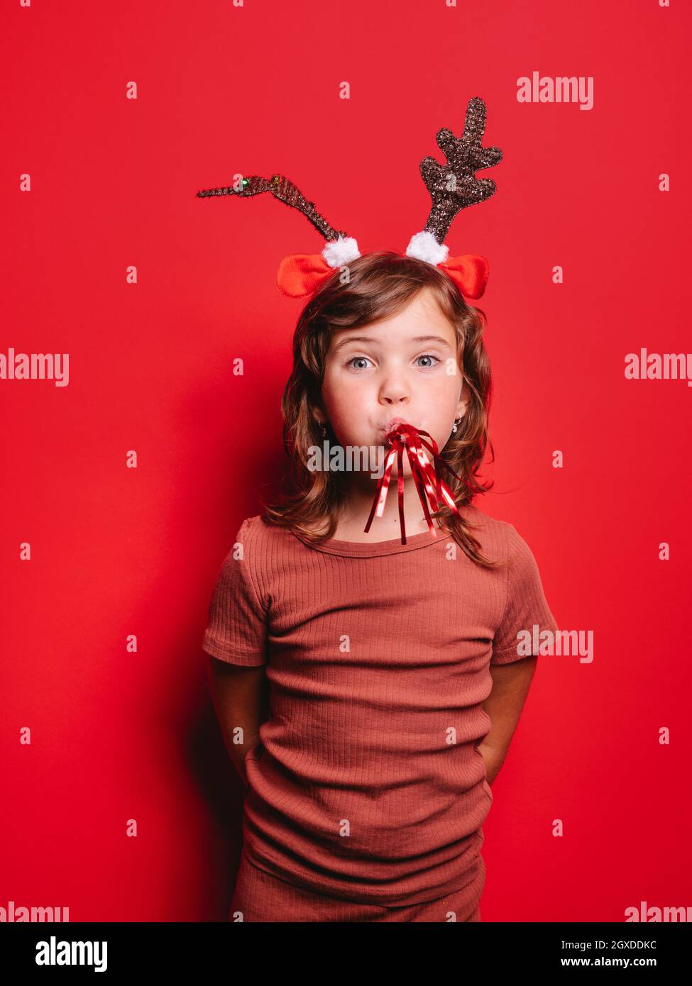 Joyful little girl in casual clothes and festive deer headband blowing party whistle and looking at camera during Christmas celebration against red ba Stock Photo