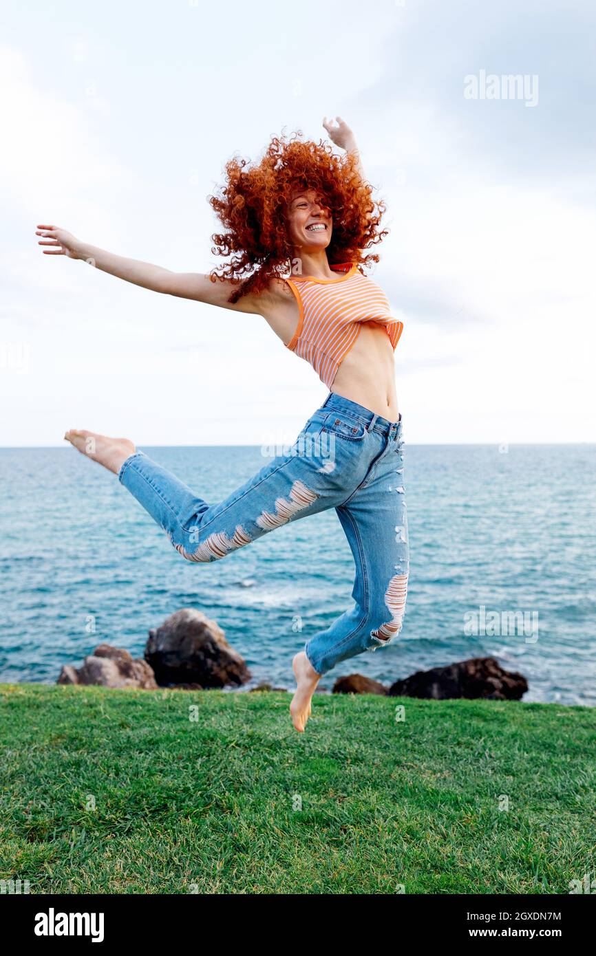 Full body of happy energetic female with flying ginger curly hair jumping  above grassy ground on coast of blue rippling sea looking away Stock Photo  - Alamy