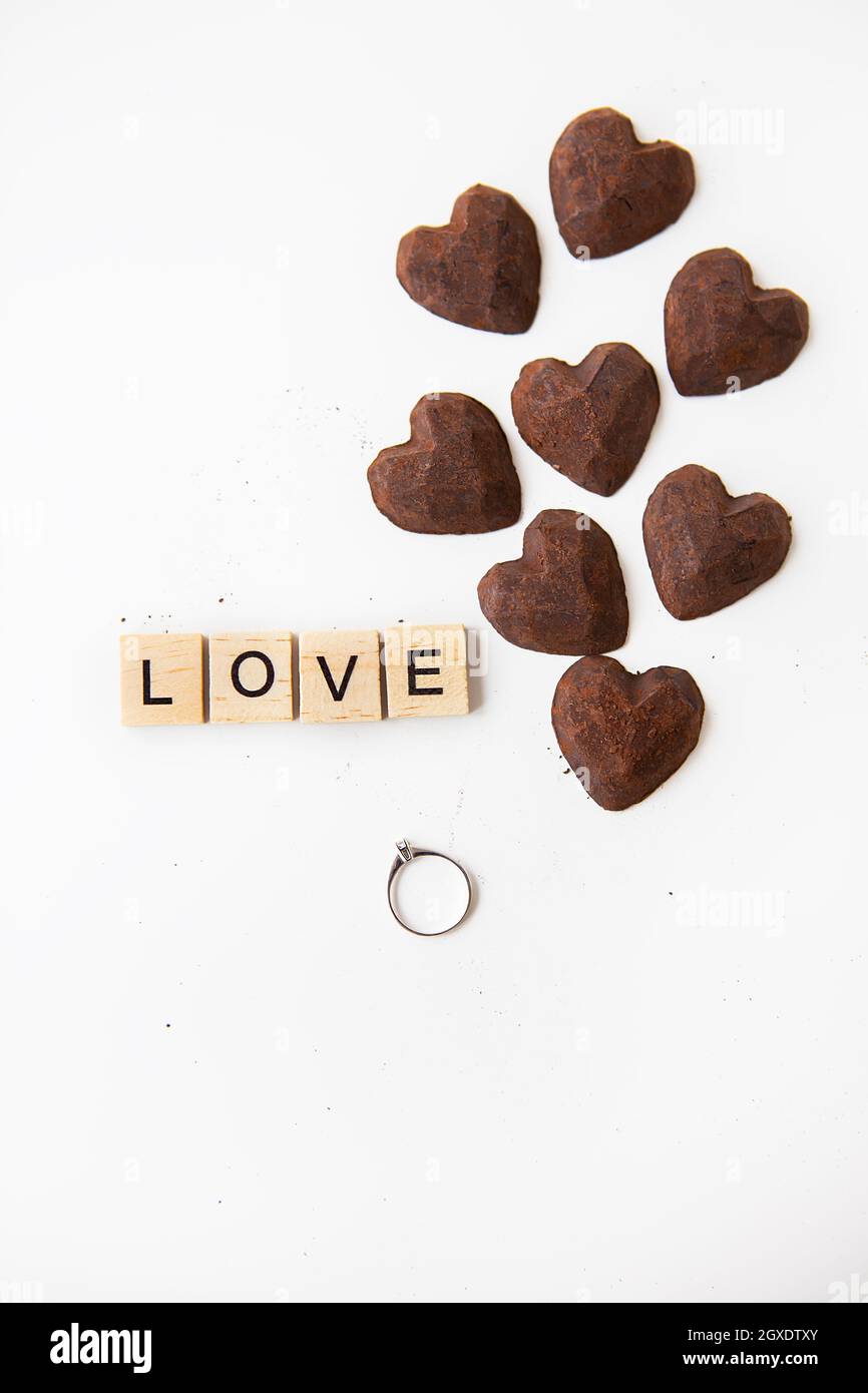 Truffle chocolate candies in the form of a heart on a white background. Inscription love made of wooden letters and a ring with a diamond. Hand and se Stock Photo