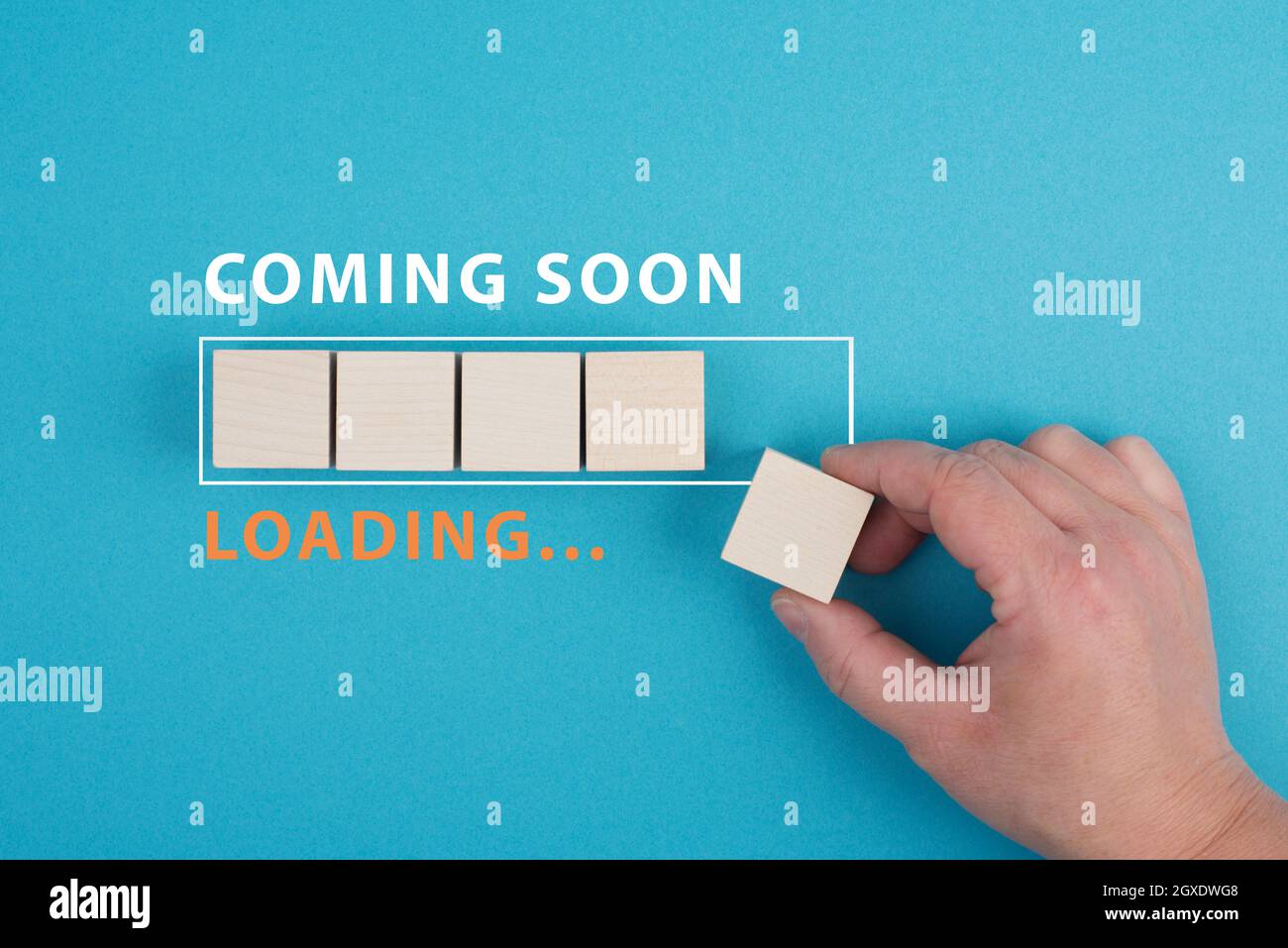The words coming soon are standing over a loading bar, hand puts the last wooden cube to the line, blue colored background Stock Photo