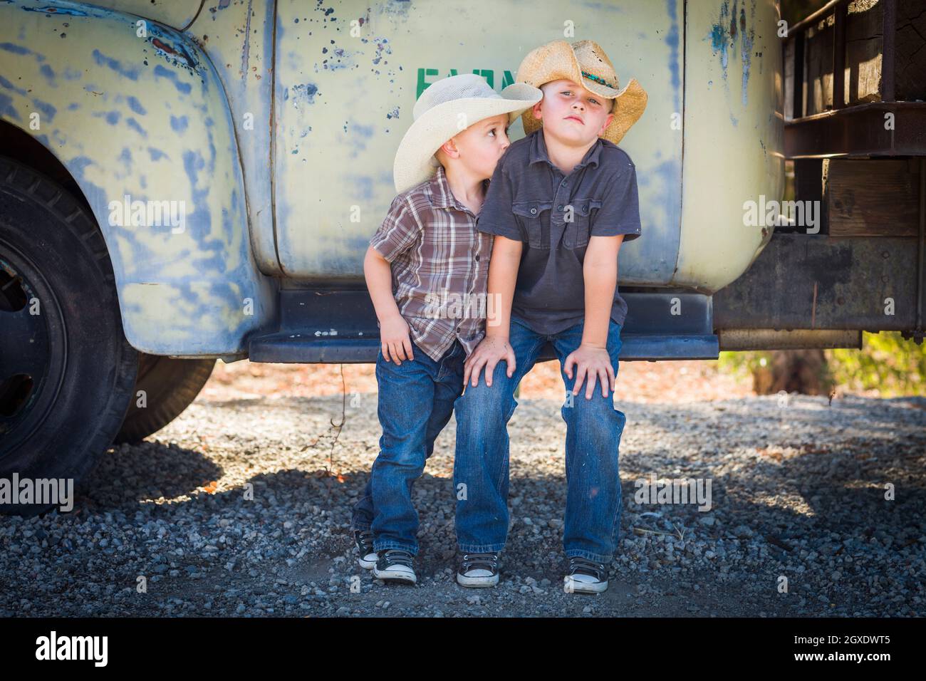 Two Young Boys Wearing Cowboy Hats Leaning Against an Antique Truck in a Rustic Country Setting. Stock Photo