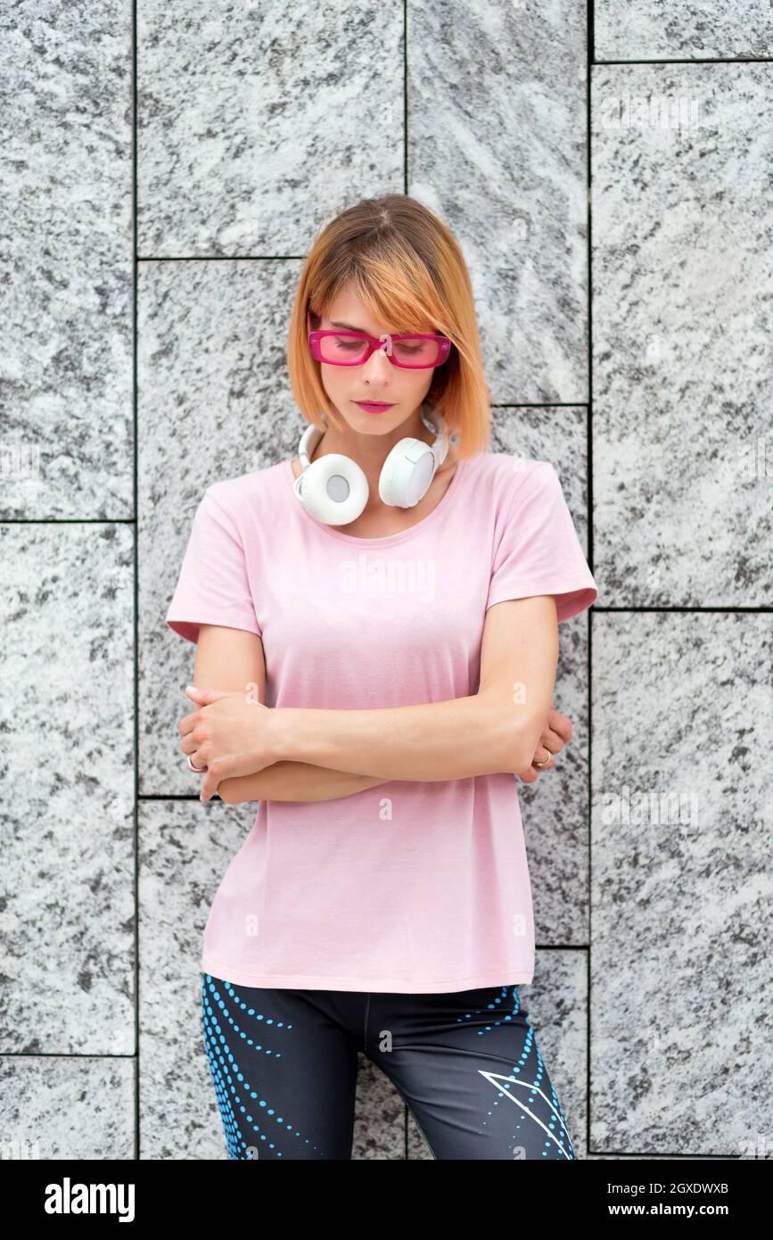 Thoughtful young female in stylish pink sunglasses with headphones on neck dressed in sporty t shirt and pants standing with arms crossed against gray Stock Photo