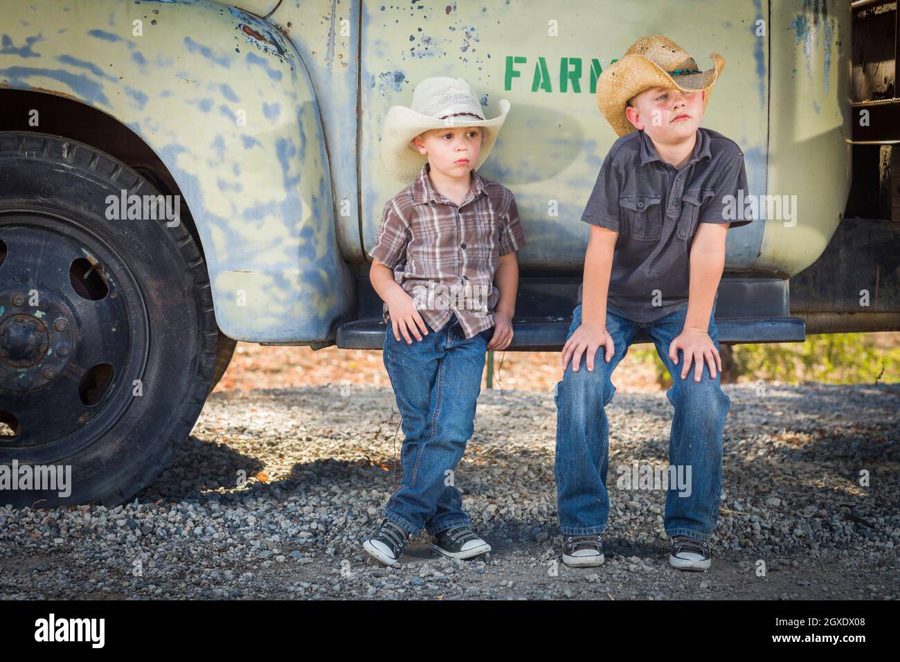 Two Young Boys Wearing Cowboy Hats Leaning Against an Antique Truck in a Rustic Country Setting. Stock Photo