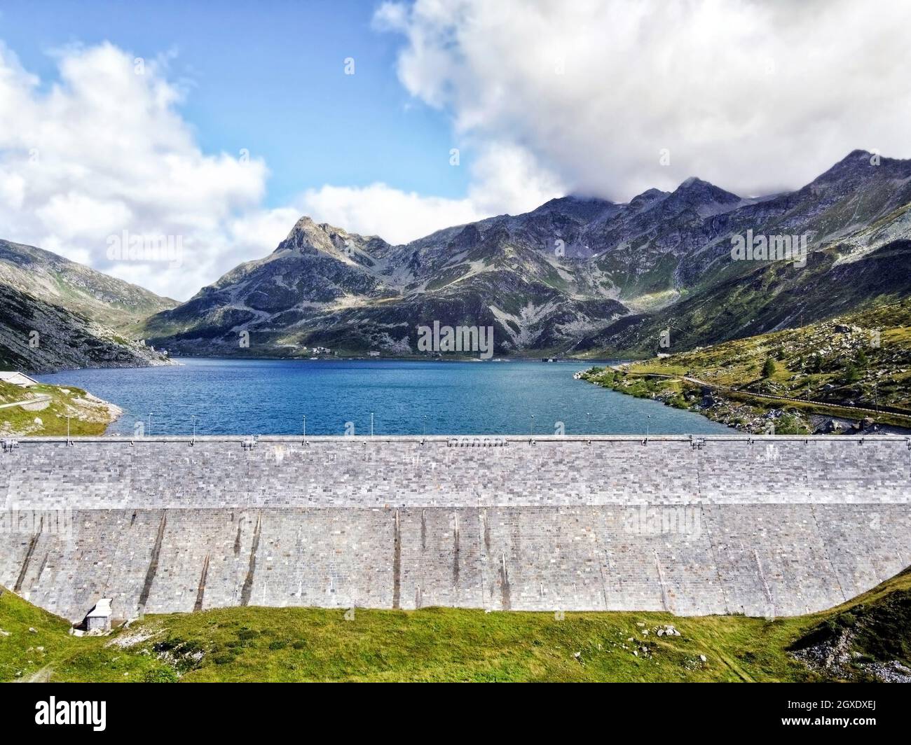 Concrete dam and water reservoir surrounded by mountains with green slopes under blue cloudy sky in summer day Stock Photo
