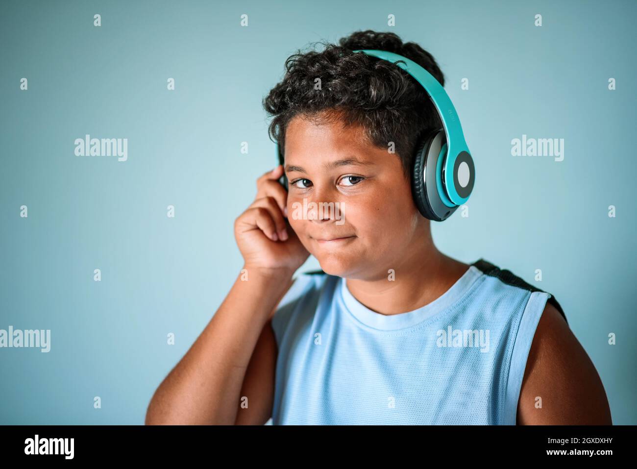 Happy smart curly haired preteen African American boy enjoying favorite music through blue wireless headphones and looking at camera in studio Stock Photo