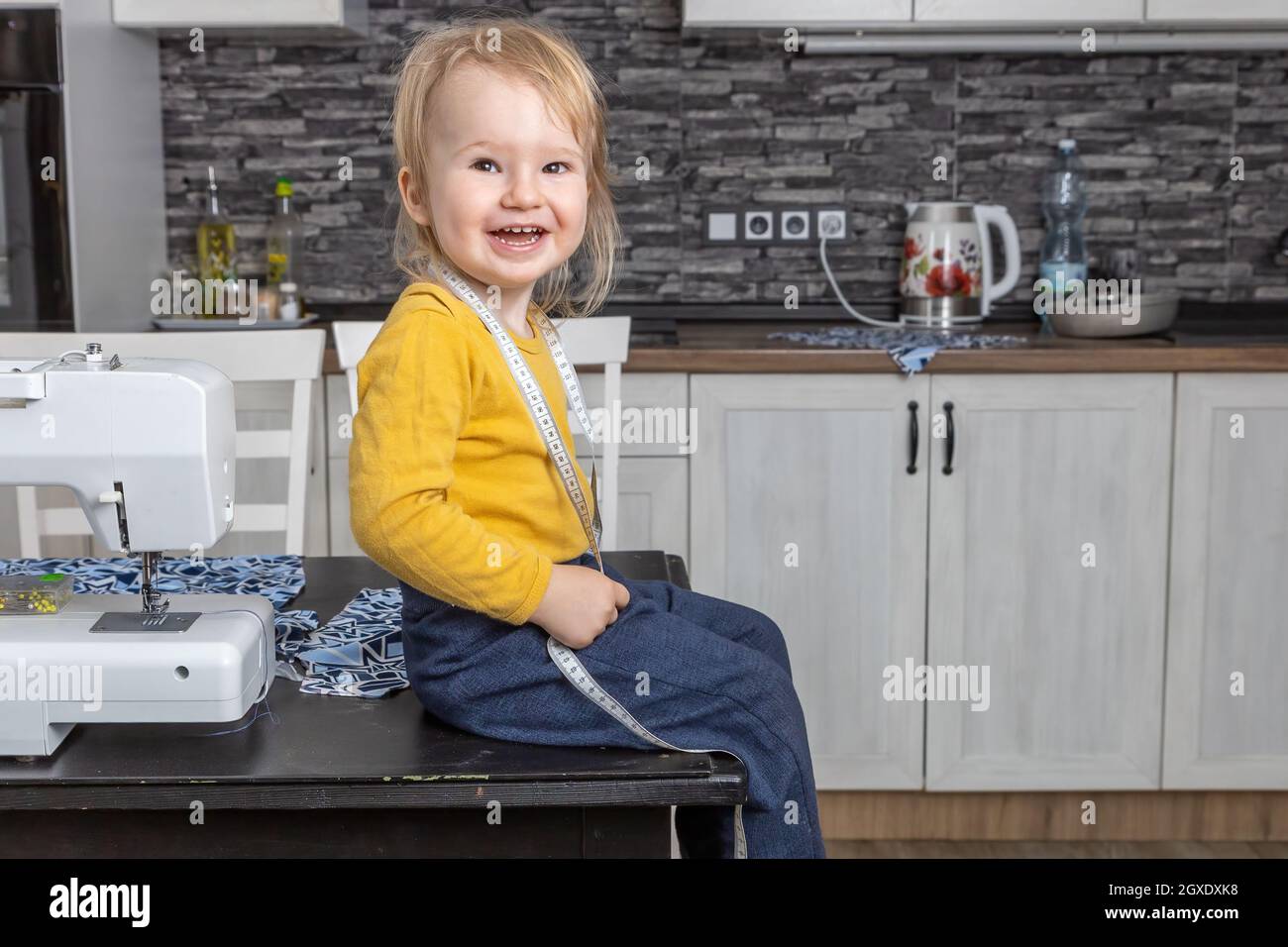 Cute laughing little boy is sitting on the table with tailor’s tape measure  smiling at the camera. Stock Photo
