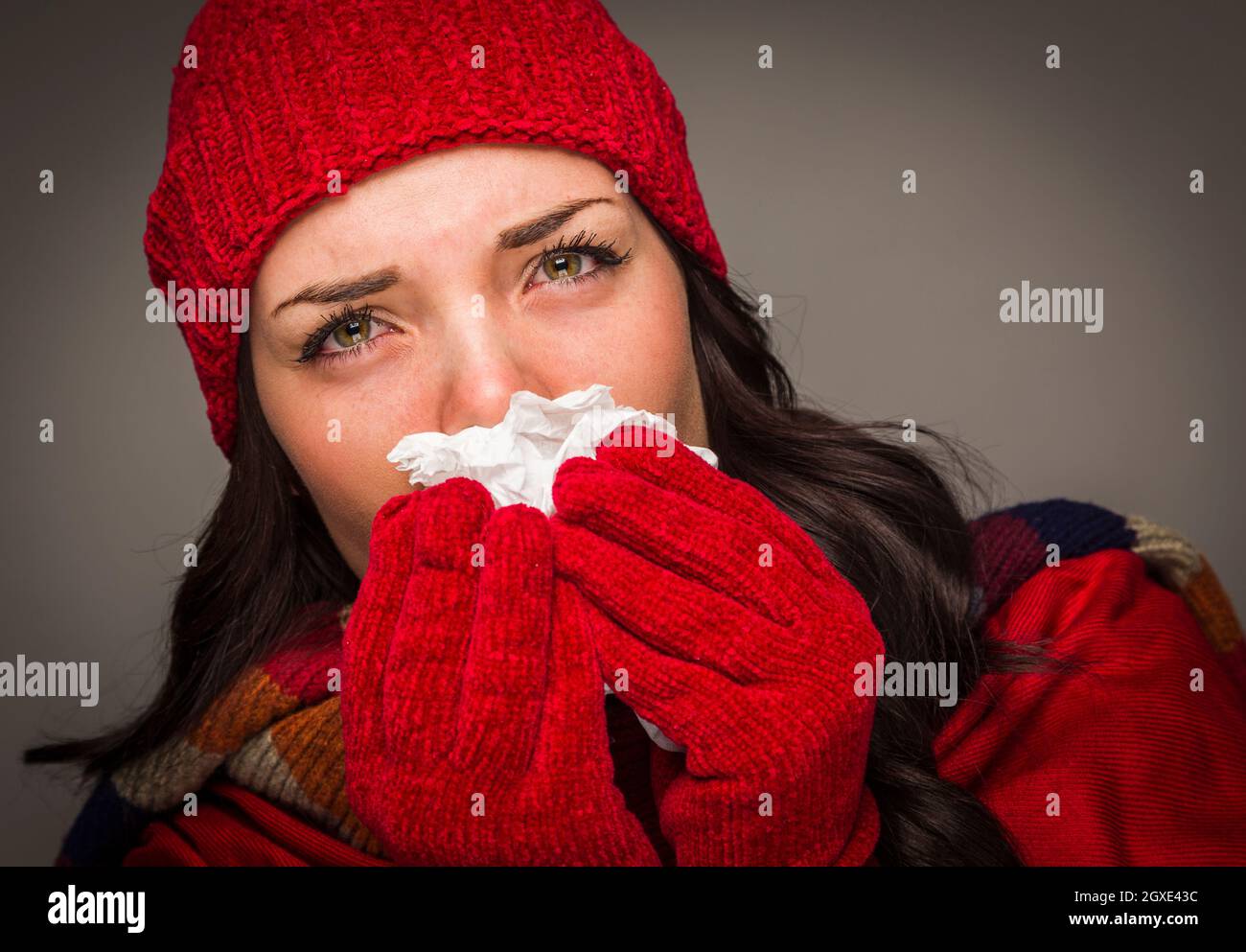 Sick Mixed Race Woman Wearing Winter Hat and Gloves Blowing Her Sore Nose with a Tissue. Stock Photo