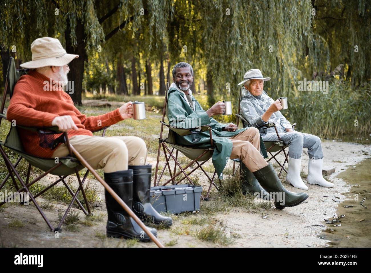 Smiling multicultural senior men in fishing outfit with thermo cups sitting  near lake in park Stock Photo - Alamy