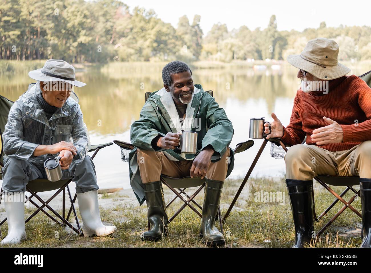 Smiling multiethnic men in fishing outfit holding thermo cups while talking  on chairs near lake Stock Photo - Alamy