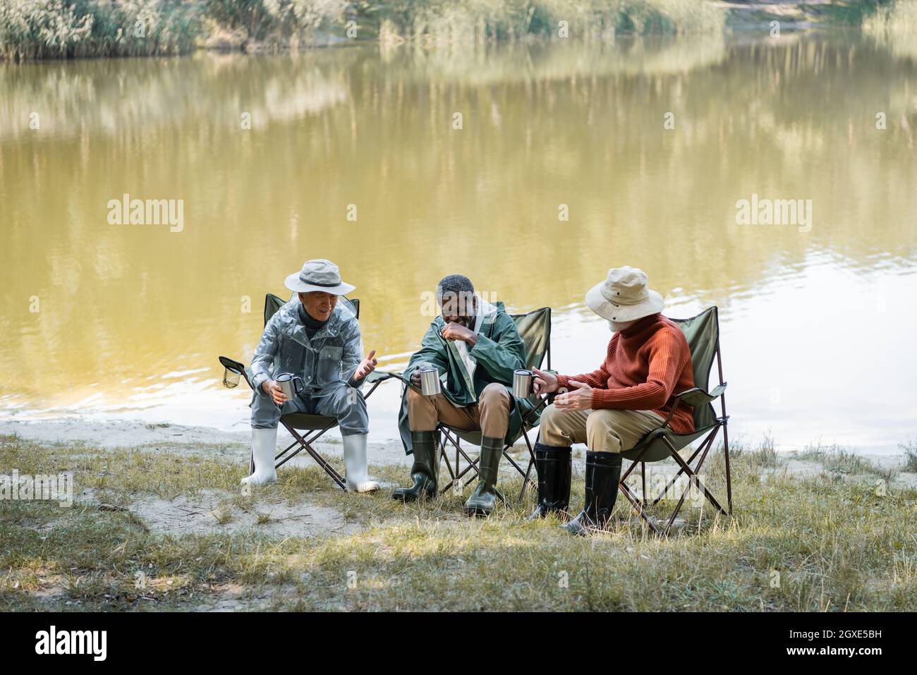 Positive interracial senior men in fishing outfit holding thermo cups while  spending time outdoors Stock Photo - Alamy