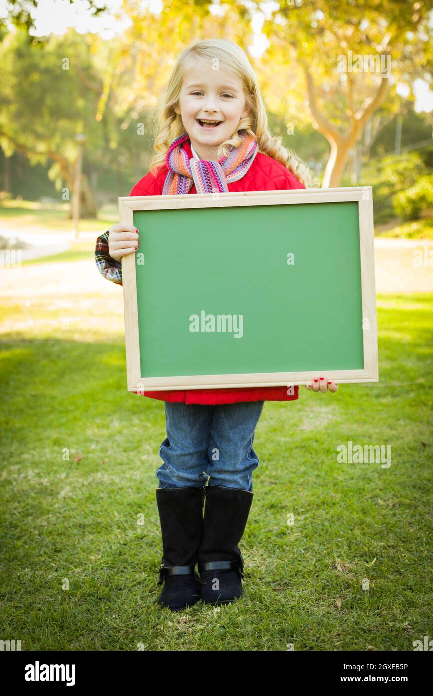 Cute Little Blonde Wearing Winter Coat and Scarf Holding a Green Chalkboard Outdoors. Stock Photo