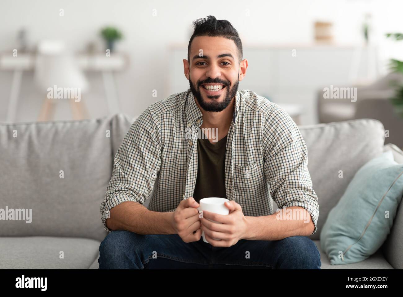 Young smiling Arab guy drinking hot aromatic coffee on sofa at home, copy space Stock Photo