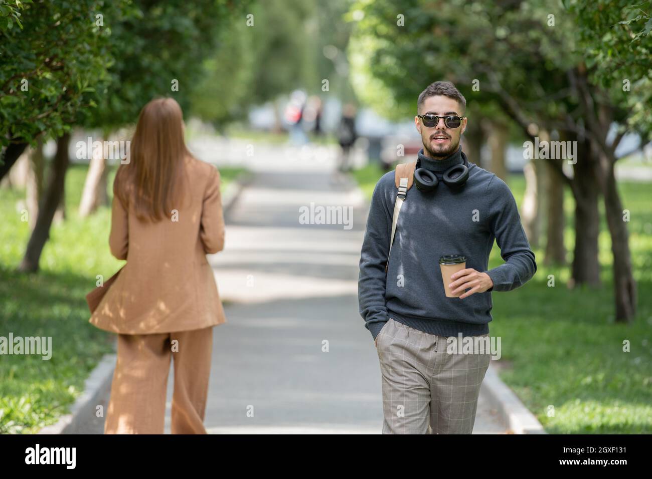 Handsome guy with drink moving down road in park with young female in suit behind Stock Photo