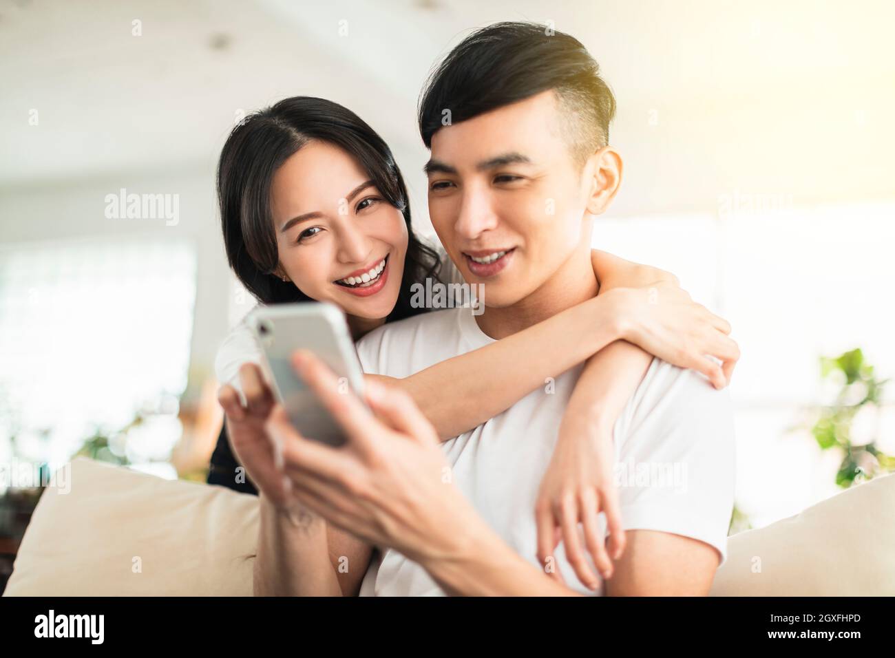 Happy young couple embracing while looking at mobile phone in living room at home Stock Photo