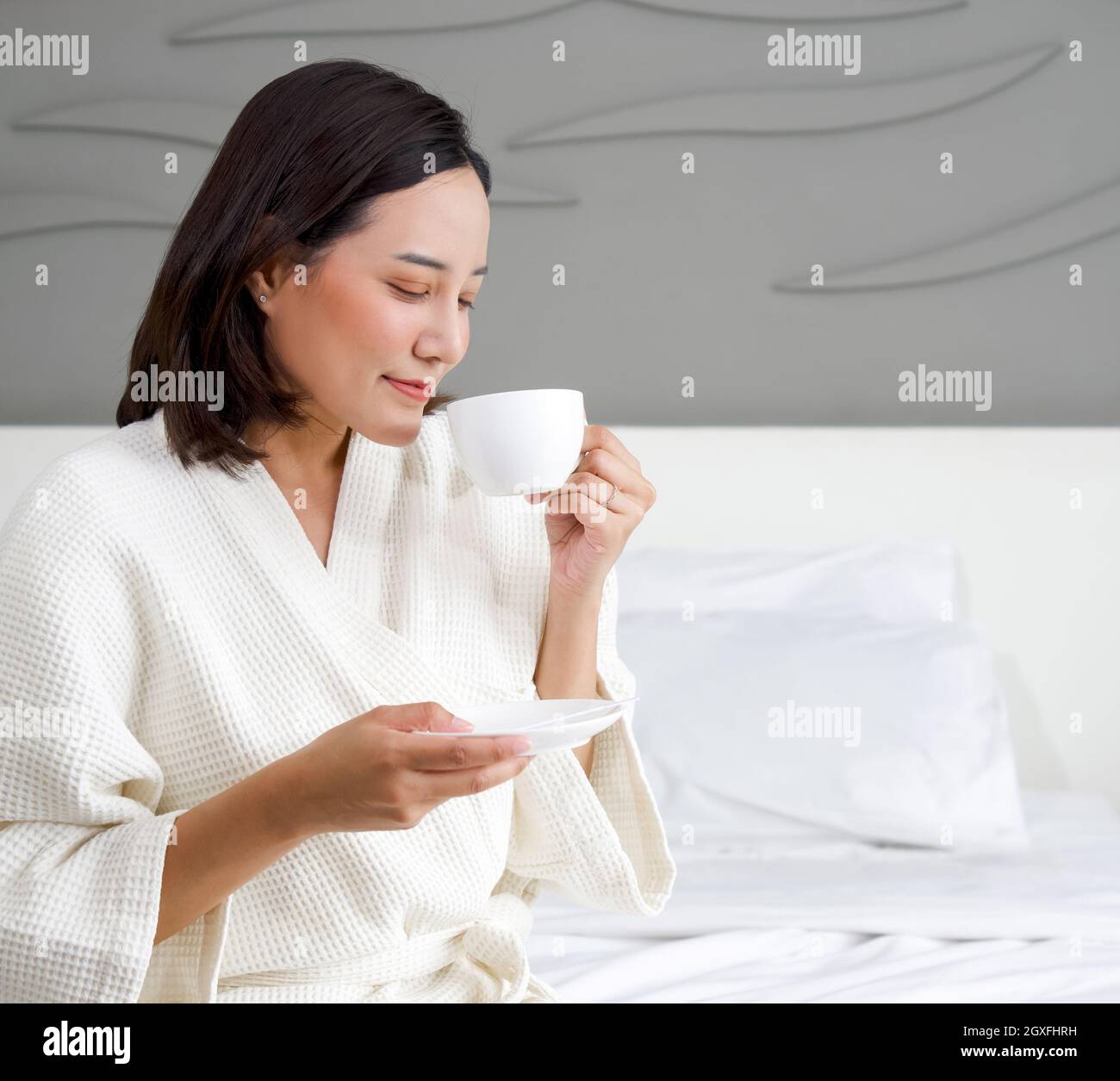 Asian woman in a bathrobe enjoys having morning tea on bed. Stock Photo