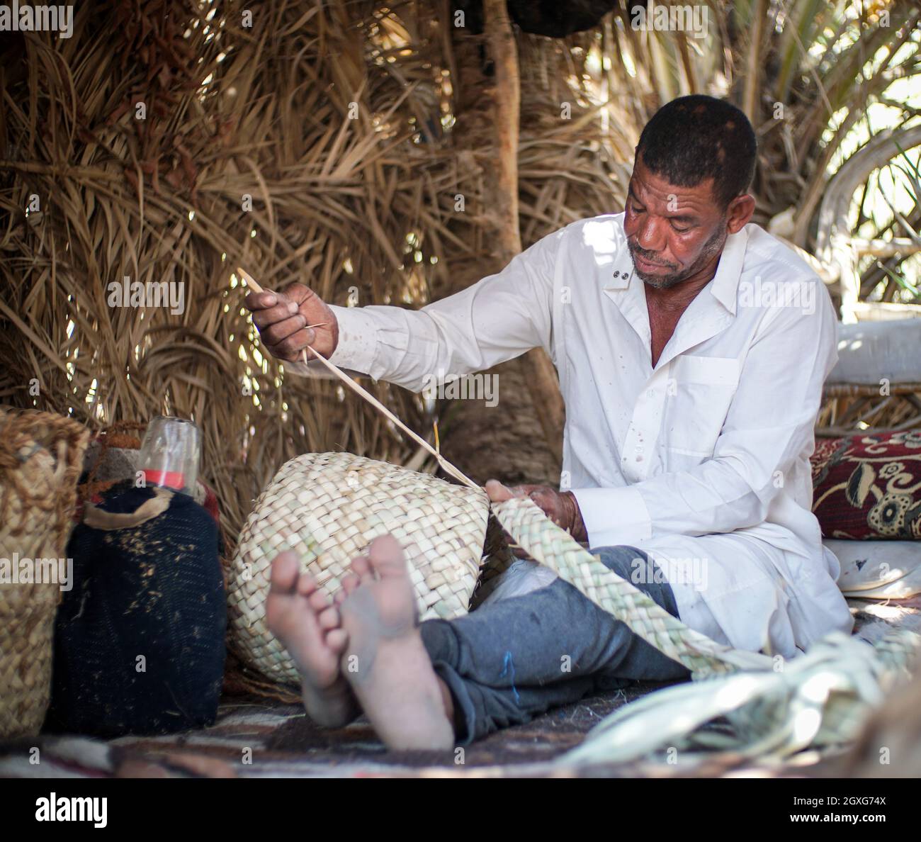 A Siwan man is weaving palm leaf into traditional bags and mat. The traditional straw plaited bags are still used widely in Siwa Oasis. Stock Photo