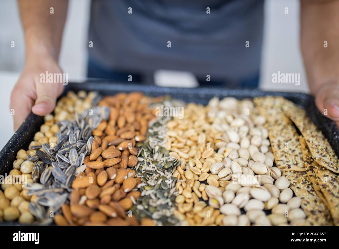 Male hands holding black tray of chickpeas, almonds, peanuts, pistachios, and multi-seed crispbread. Fresh crispy bread, seeds, and nuts. Healthy snac Stock Photo