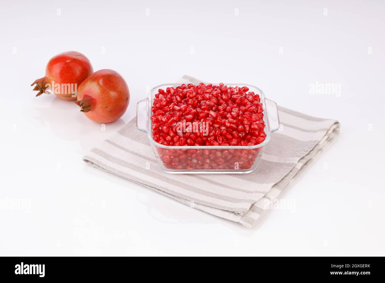 Fresh Pomegranate seed arranged in a square glass container with fresh fruit placed beside it on white background, isolated. Stock Photo
