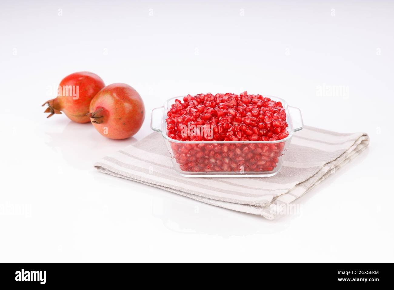 Fresh Pomegranate seed arranged in a square glass container with fresh fruit placed beside it on white background, isolated. Stock Photo