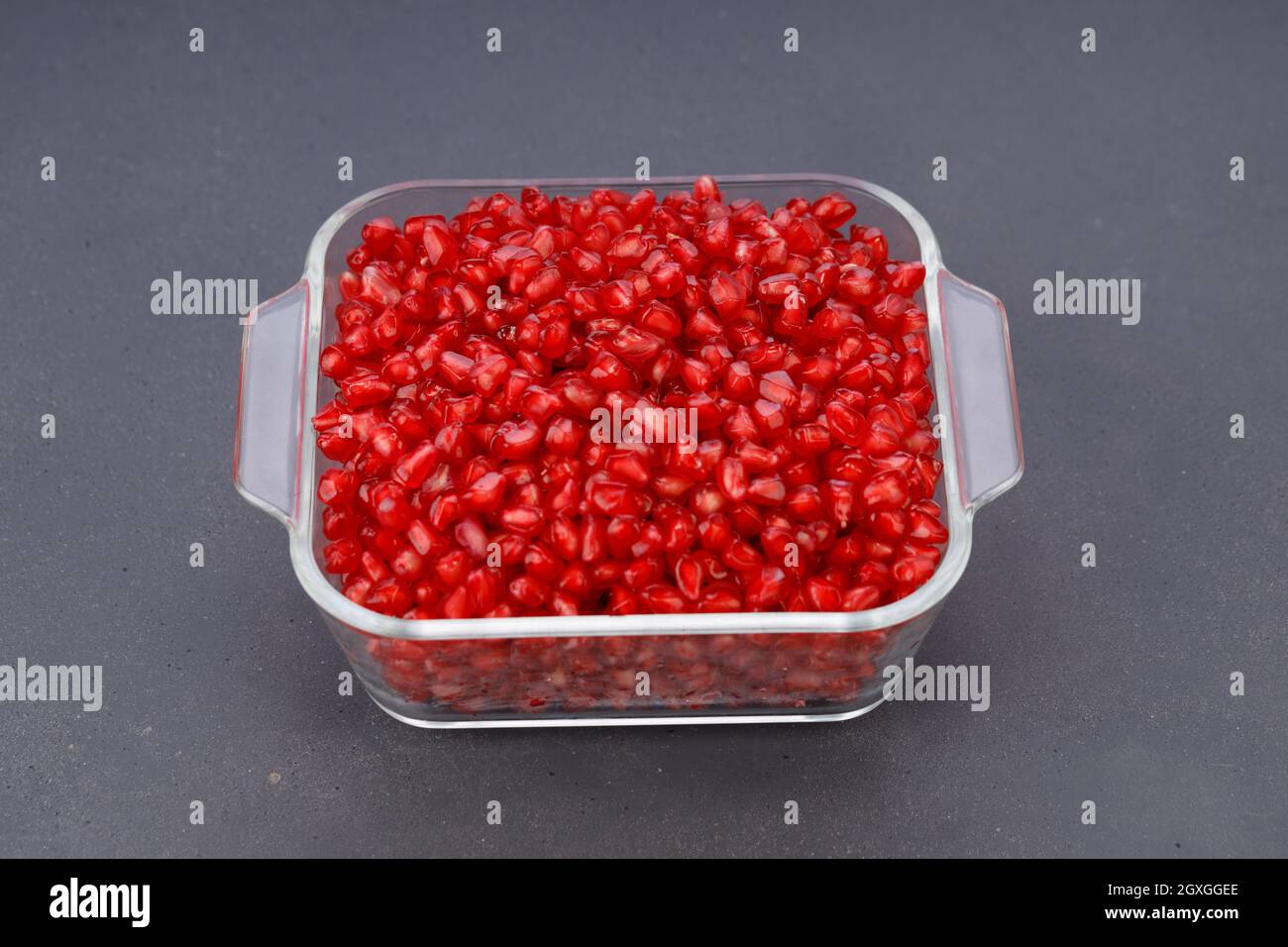 Fresh Pomegranate seed arranged in a square glass container with fresh fruit placed beside it on grey background, isolated. Stock Photo