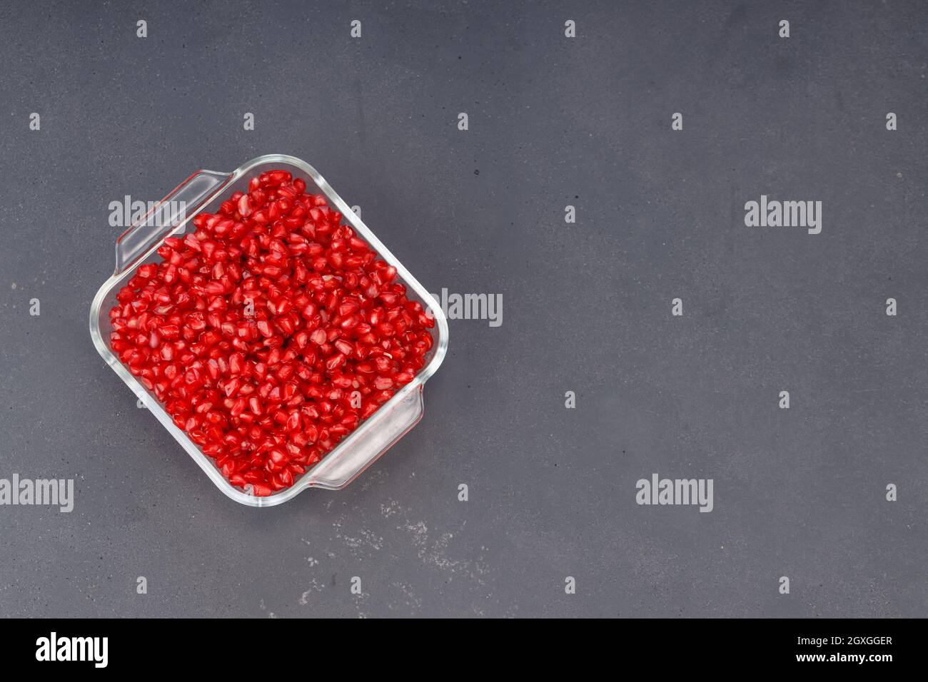 Fresh Pomegranate seed arranged in a square glass container with fresh fruit placed beside it on grey background, isolated. Stock Photo