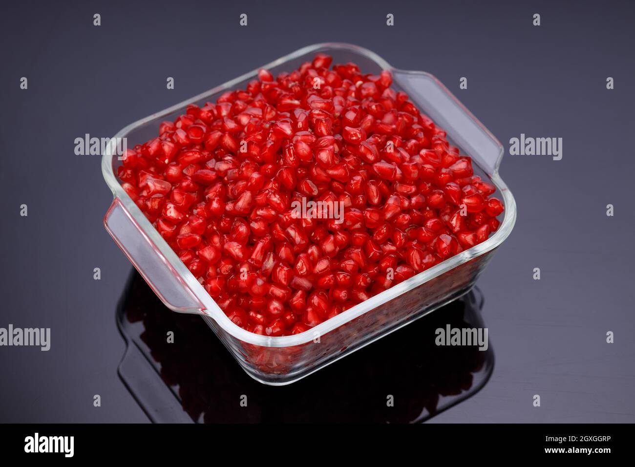 Fresh Pomegranate seed arranged in a square glass container with fresh fruit placed beside it on grey background, isolated. Stock Photo