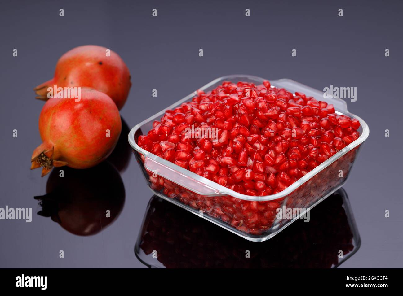 Fresh Pomegranate seed arranged in a square glass container with fresh fruit placed beside it on grey background, isolated. Stock Photo