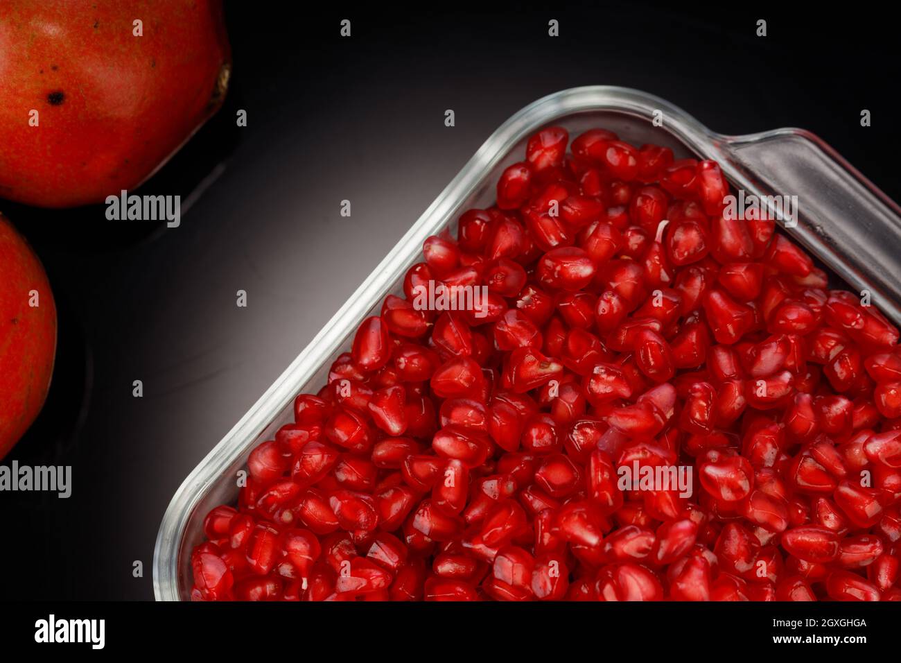 Fresh Pomegranate seed arranged in a square glass container with fresh fruit placed beside it on grey background, isolated. Stock Photo