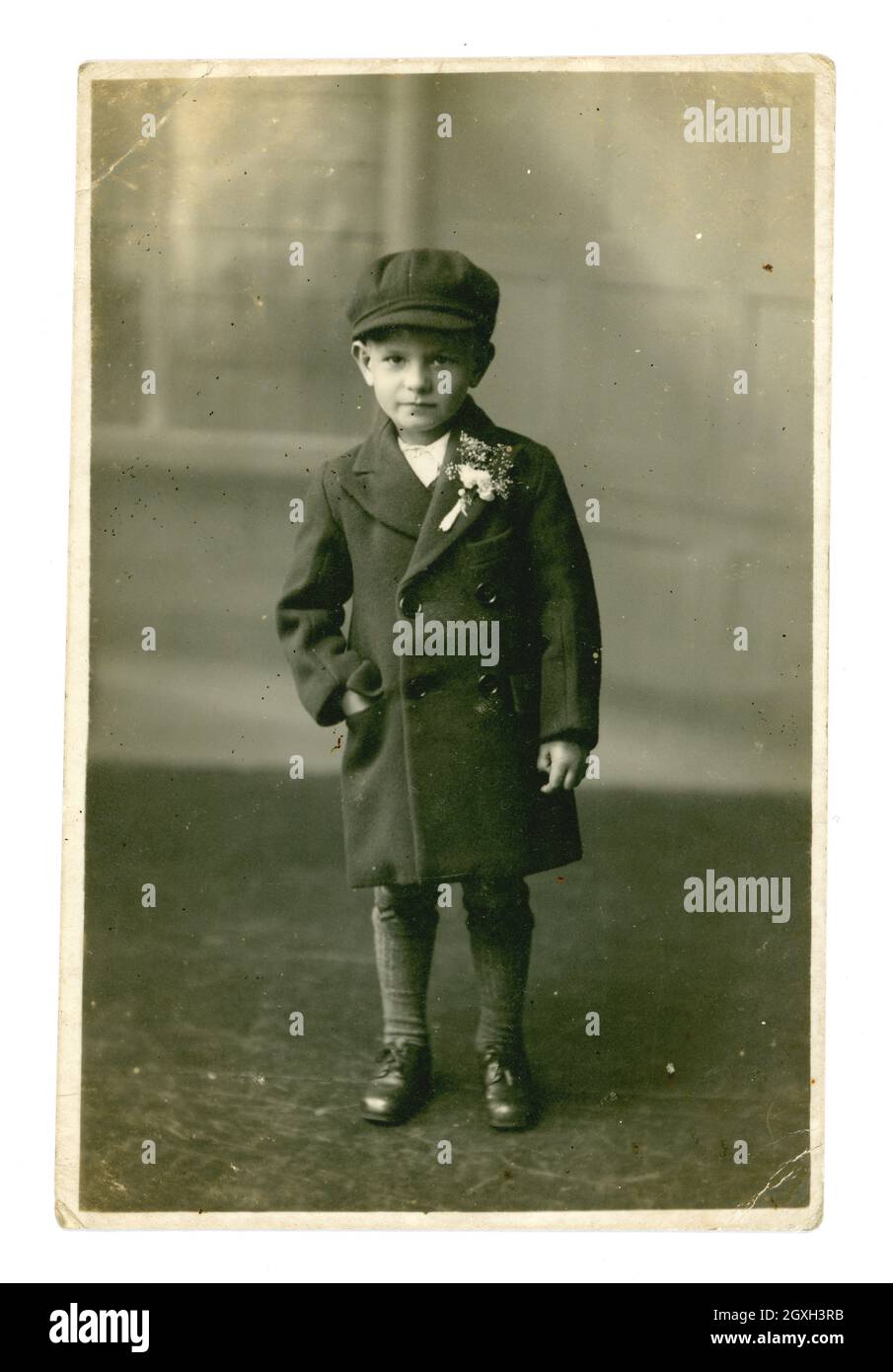 Original early 1940's postcard studio portrait of cute serious looking working class young lad wearing a smart long jacket, with buttonhole flowers on his lapel, and a flat cap. 1940's boy. 1940's child. 1940's children. He is maybe a pageboy at a wedding, Cheriton, Folkestone, Kent, U.K. dated July 6 1940 Stock Photo