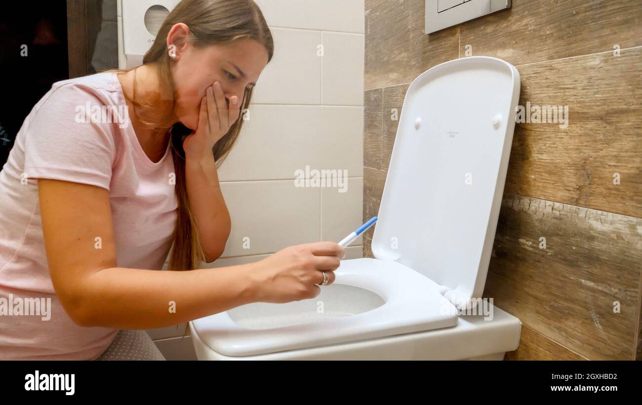 Young woman looking on positive pregnancy test sitting on toilet and