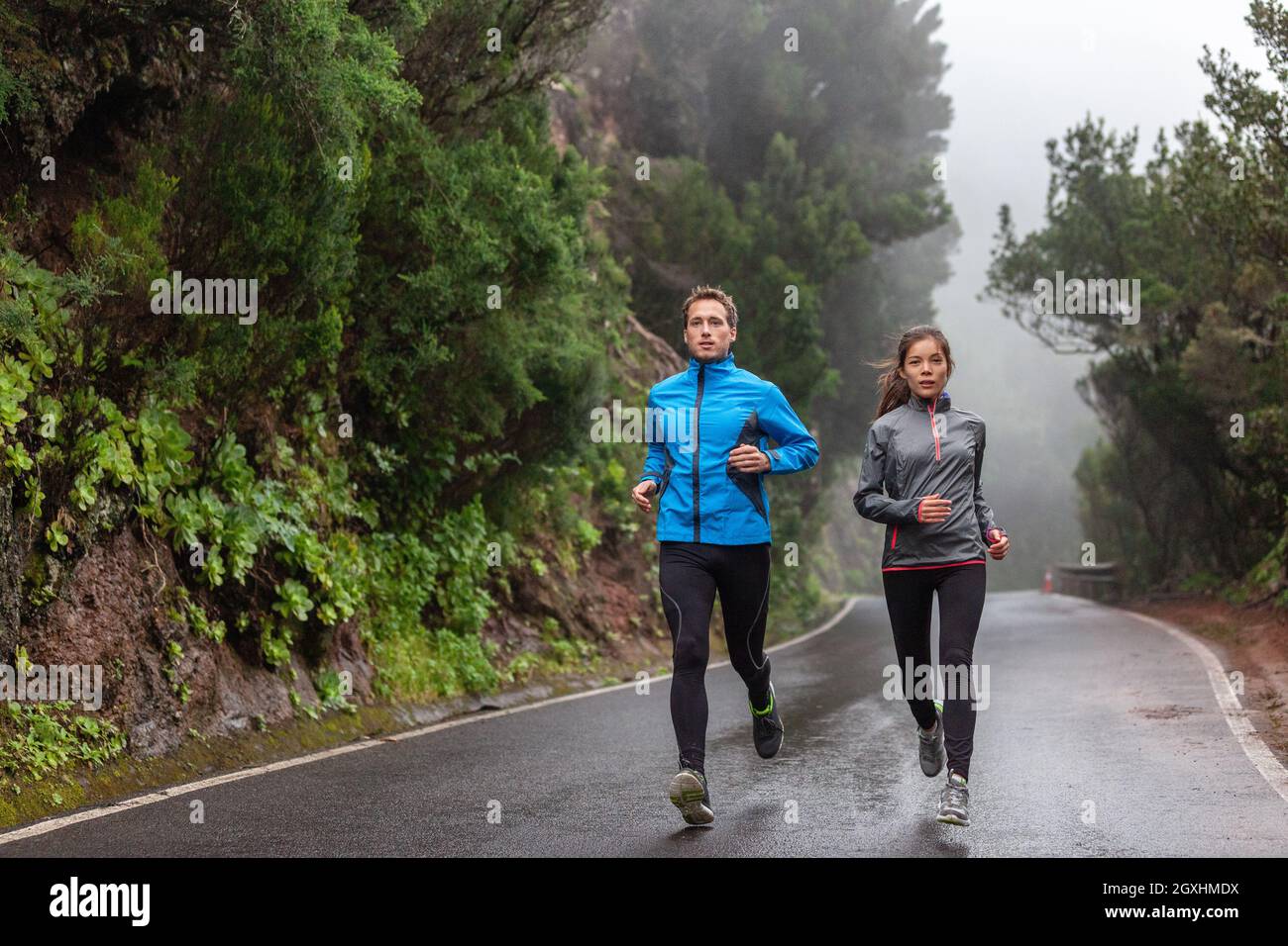 Rain run fit people training jogging in rain weather wearing cold clothing running outdoors in nature autumn season. Active couple on wet park trail Stock Photo