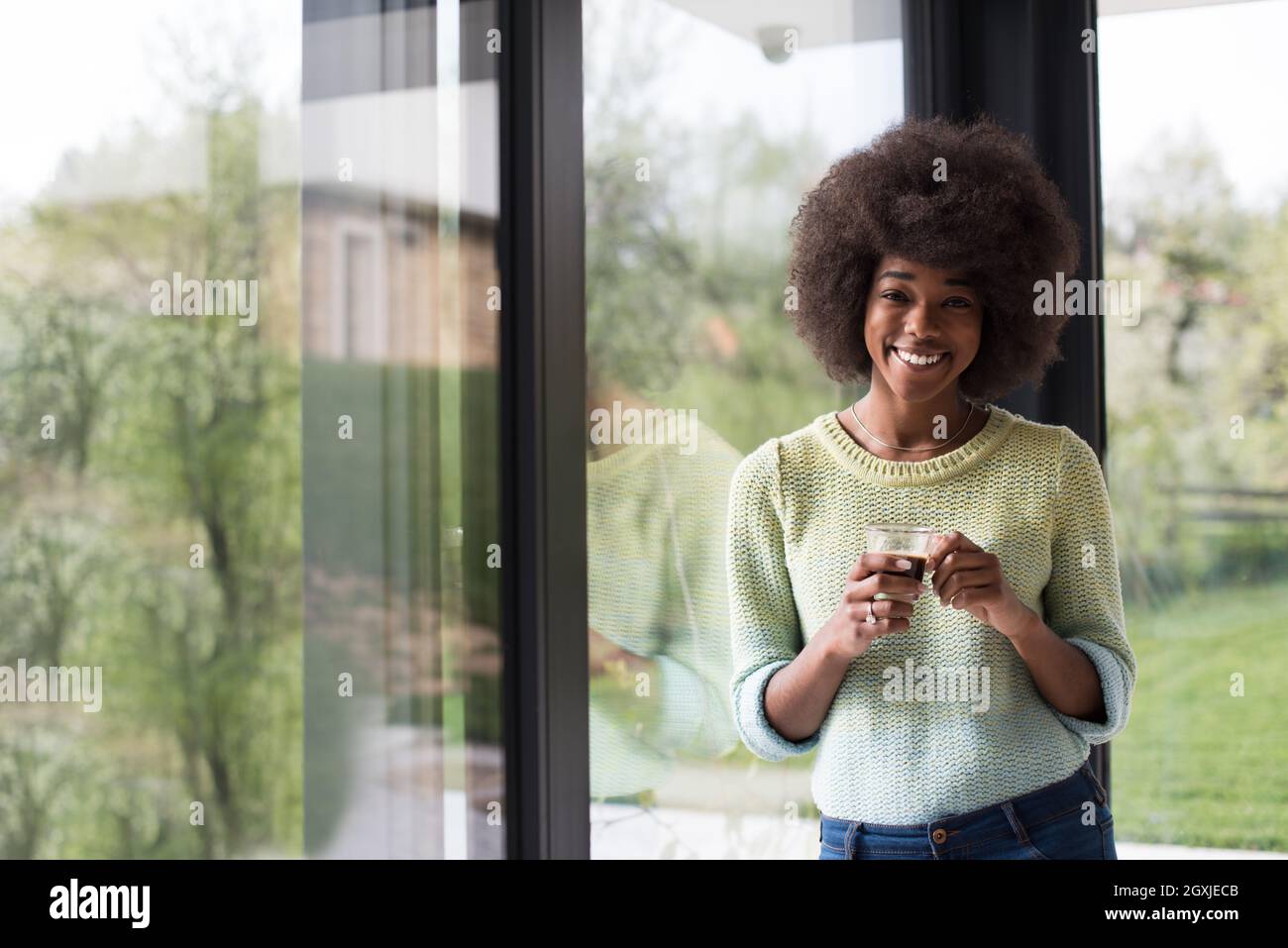 Beautiful young african american woman drinking coffee and looking through a window in her luxury home Stock Photo