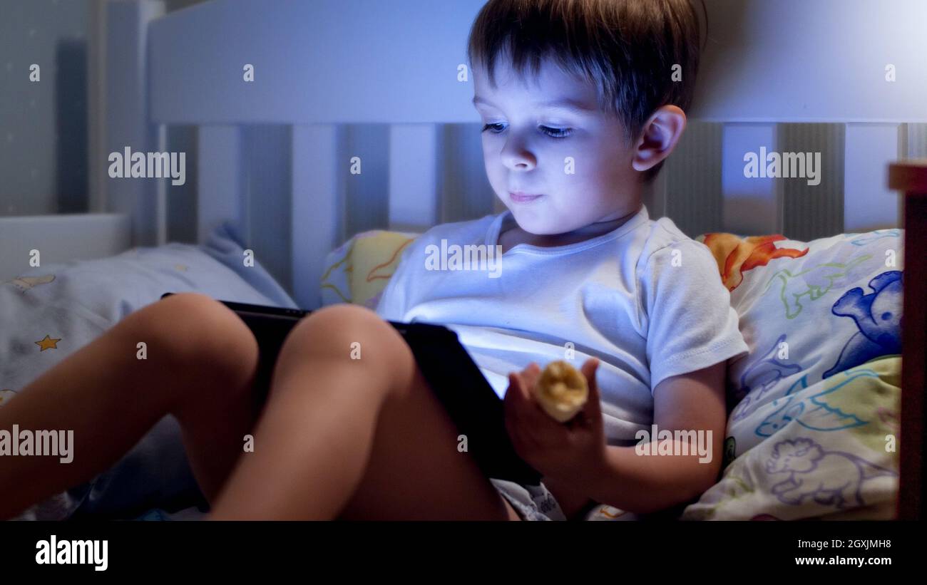 Portrait of cute little boy lying in bed at night and using digital ...