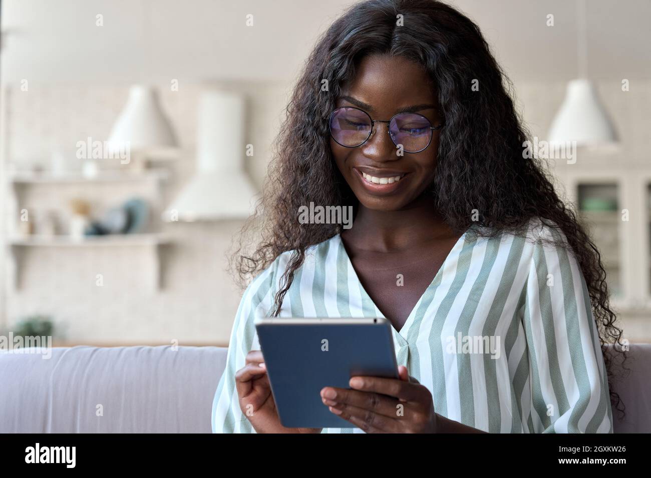 Smiling happy black girl in glasses sits on sofa at home holding tablet device. Stock Photo