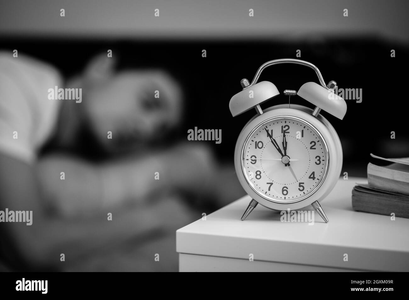 White alarm clock on the bedside table. Young man sleeps in the background. Stock Photo