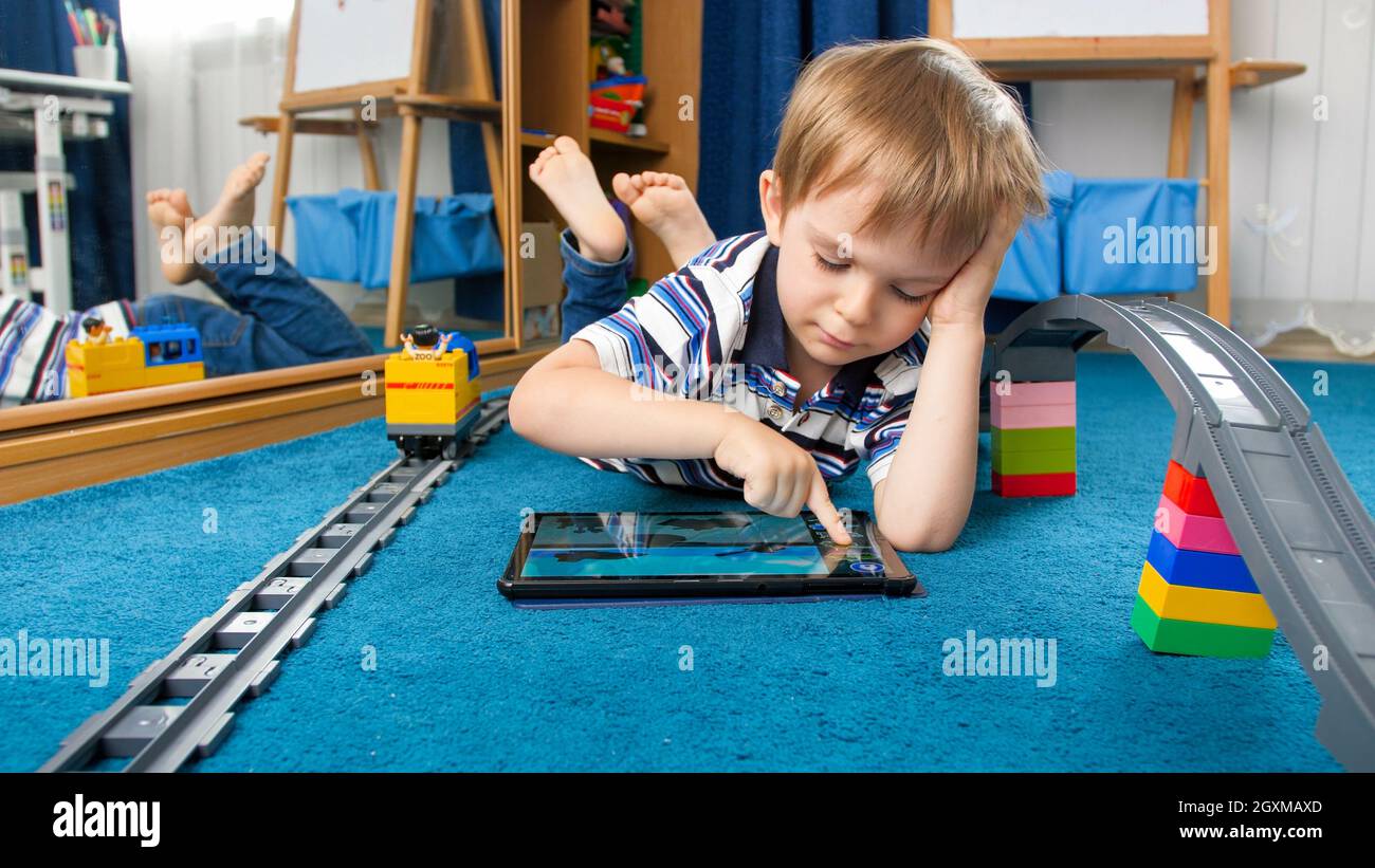 Cute little boy lying on floor and playing educational games on tablet ...