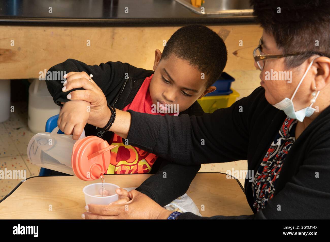 Education Preschool 4-5 year olds female teacher assists boy pouring water at meal time Stock Photo