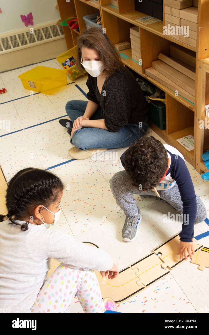 Education Preschool 4-5 year olds female intern graduate student observes play between a boy and a girl in the block area, wearing face masks Covid-19 Stock Photo