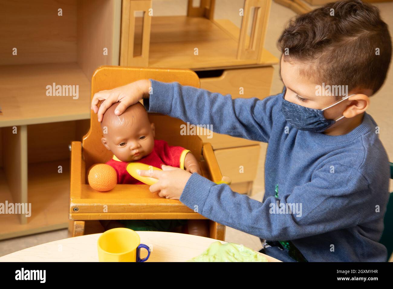 Education Preschool 3-4 year olds pretend play in family area, boy feeding doll with play food, wearing face mask to protect against Covid-19 Stock Photo