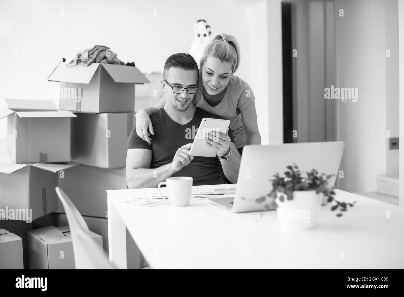 Young couple moving in a new home. Man and woman at the table using notebook laptop computer and plans with boxes around them Stock Photo