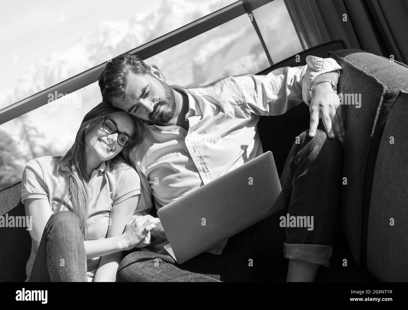 Young couple relaxing at  home using laptop computers reading in the living room near the window on the sofa couch. Stock Photo